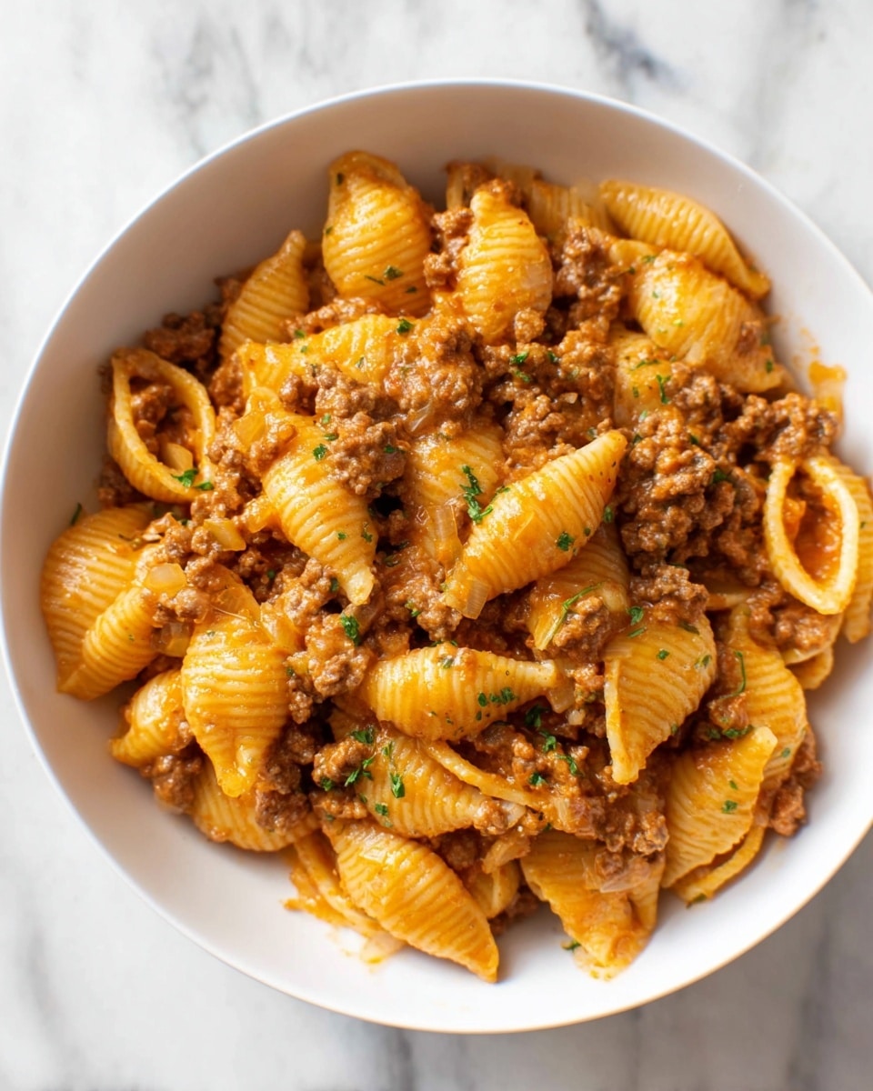 A white bowl filled with pasta shells mixed with ground meat and a lightly orange sauce. The pasta shells are firm and ridged, coated evenly in the sauce with small bits of cooked onion scattered throughout. The ground meat is browned and crumbly, blended well with the sauce and pasta. Small green herb pieces are sprinkled on top, adding a touch of color. The bowl sits on a white marbled surface. photo taken with an iphone --ar 4:5 --v 7