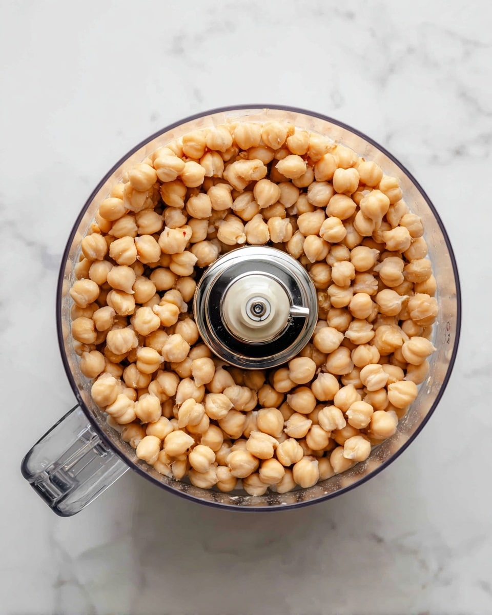 A clear food processor bowl filled with a single layer of light beige chickpeas. The bowl is round with a handle on the left side, and inside the bowl is a silver blade with a black center in the middle, surrounded by the chickpeas. The food processor is placed on a white marbled surface. photo taken with an iphone --ar 4:5 --v 7