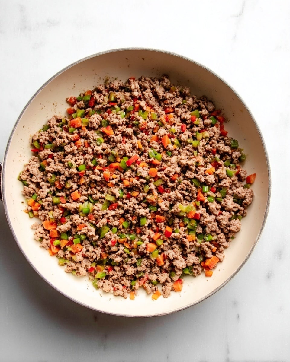 The image shows a round white pan filled with cooked ground meat mixed with small pieces of red and green vegetables, spread evenly across the pan. The meat has a slightly browned, crumbly texture, while the vegetables add pops of bright red and green colors throughout. The pan sits on a white marbled surface, giving a clean and simple background that makes the food stand out. photo taken with an iphone --ar 4:5 --v 7