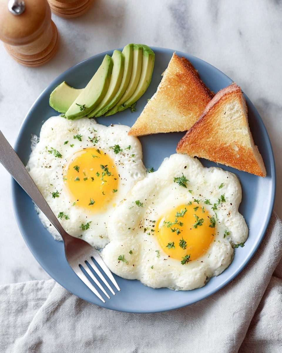 A white plate with two cloud-like eggs that have fluffy white edges and bright yellow yolks in the center, sprinkled with small green herbs; to the left of the eggs are three light green slices of avocado, and on the right side, there are two triangular slices of toasted bread with a golden-brown color. A silver fork is on the plate near the toast, and the plate is on a white marbled surface with a light gray cloth and a wooden pepper shaker nearby. photo taken with an iphone --ar 4:5 --v 7