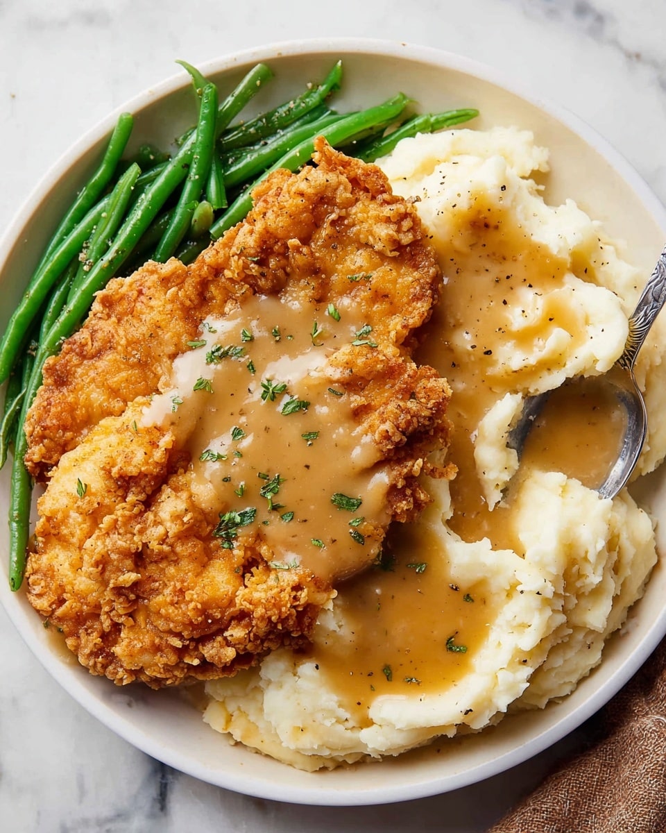 A white round plate on a white marbled surface holds a meal with three main parts: a serving of green beans on the left side, two large pieces of crispy golden-brown fried chicken layered in the center, and a generous portion of creamy mashed potatoes on the right. The mashed potatoes have smooth, fluffy texture with a spoon resting partially buried in them. Both the fried chicken and mashed potatoes are covered with a thick, light brown gravy drizzled evenly on top, sprinkled lightly with green herbs and black pepper. Photo taken with an iphone --ar 4:5 --v 7