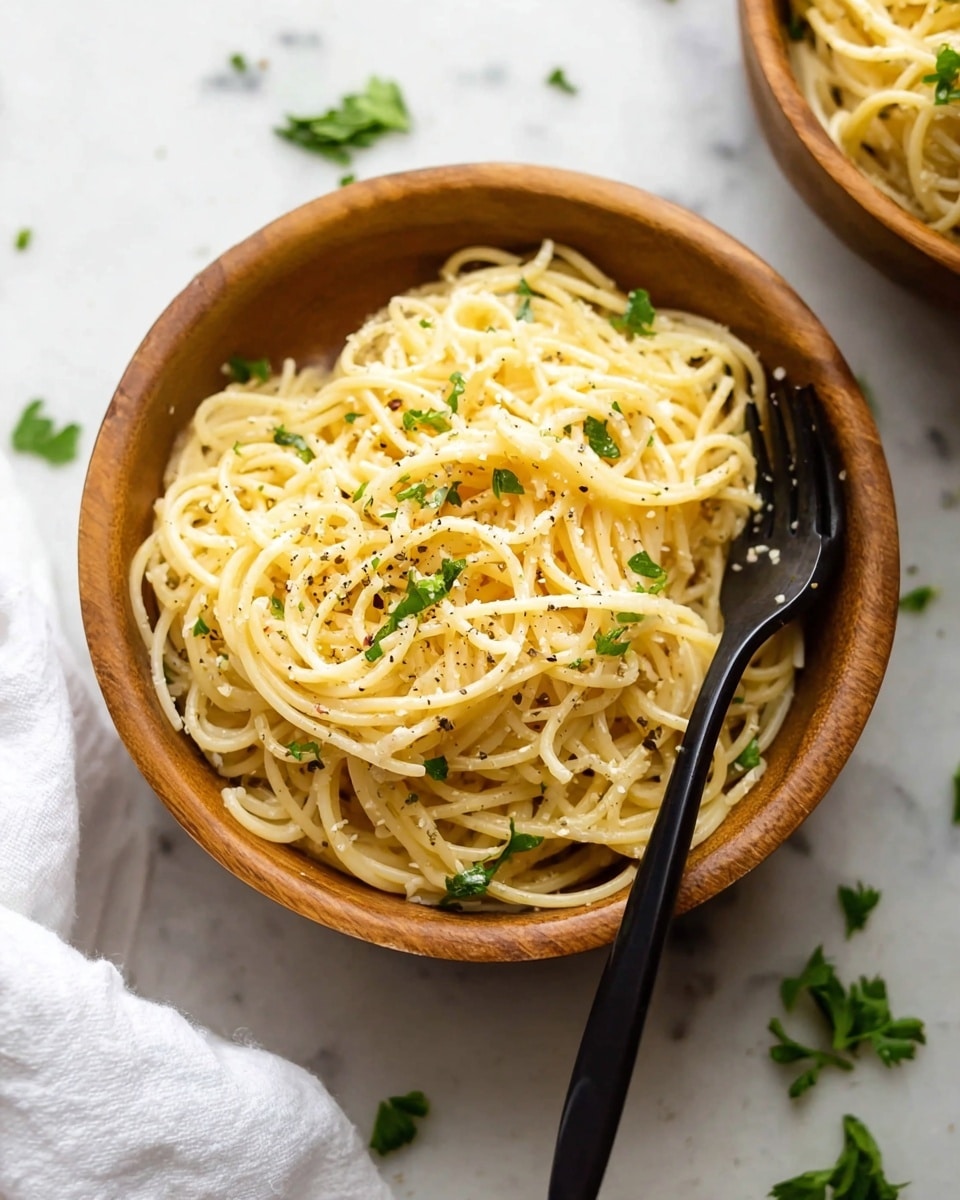 A round wooden bowl filled with thin spaghetti noodles layered loosely with light yellow color, sprinkled with small green parsley leaves and small black pepper dots spread evenly on the noodles. A black fork rests on the right side, with its tines partially stuck into the noodles. In the background, the corner of a second wooden bowl filled with spaghetti is partly visible. The scene is set on a white marbled surface with scattered parsley leaves and a white cloth napkin on the lower left side. Photo taken with an iphone --ar 4:5 --v 7