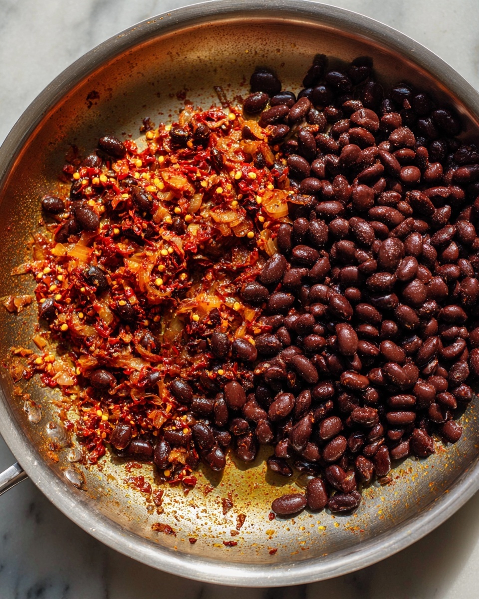 A large stainless steel pan shows one layer of cooked black beans, dark brown and smooth in texture, mostly on the right side. The left side holds a thick layer of sautéed onions and red chili flakes mixed with oil, having a rich orange-red color with glossy bits. The ingredients are spread unevenly in the pan, with cooked spices and oil visible on the pan's surface. The photo has warm, natural light highlighting the colors and textures of the dish on a white marbled surface. photo taken with an iphone --ar 4:5 --v 7