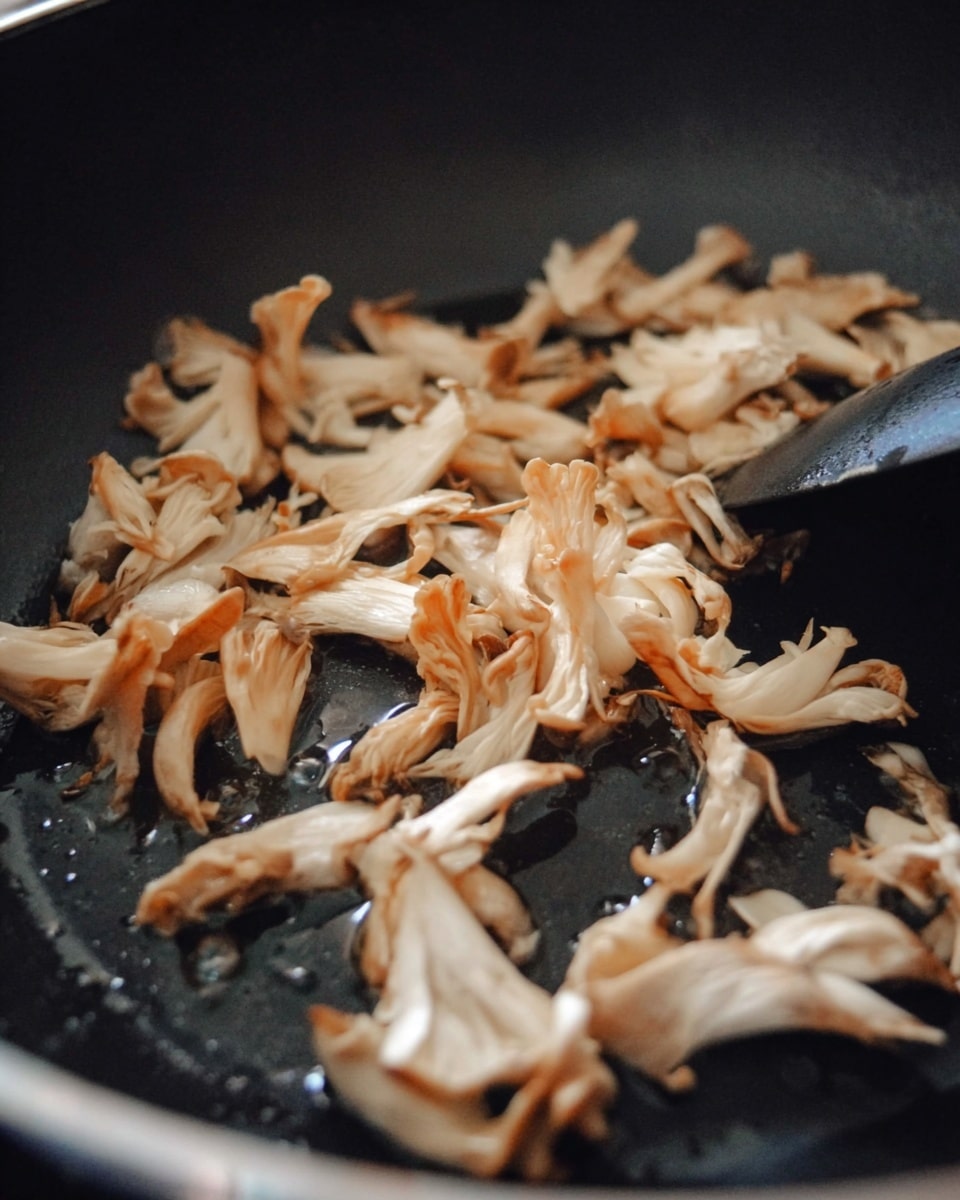 The image shows a close-up of light beige mushrooms being cooked in a black pan. The mushrooms have thin, wrinkled, and slightly curled layers with some edges turning golden brown from cooking. They are spread unevenly across the bottom of the pan, which has a shiny surface from oil. A black utensil is partially visible on the right side, stirring the mushrooms gently against the pan’s dark background. The overall colors are soft off-white with touches of golden brown on the mushrooms, contrasted by the deep black of the pan. photo taken with an iphone --ar 4:5 --v 7