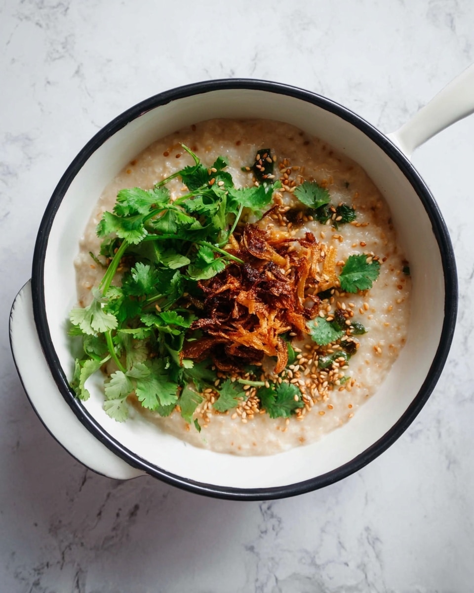 A white bowl with a black rim holds creamy beige porridge as the base layer, topped with a small pile of golden brown fried onions in the center. Fresh green cilantro leaves are scattered on and around the fried onions, adding a bright contrast. Small light brown sesame seeds are sprinkled over the dish, giving texture and detail. The bowl sits on a white marbled surface, and a white spoon with a hole at the handle rests nearby. Photo taken with an iphone --ar 4:5 --v 7