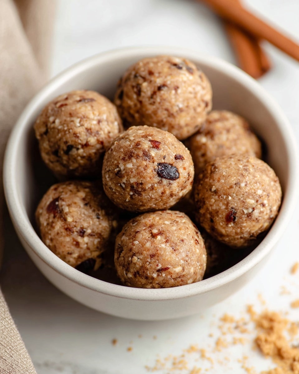 A white bowl filled with eight round energy balls that have a rough texture showing small bits of nuts and dried fruit throughout. The balls are light brown with some darker brown spots, indicating mixed ingredients like oats and dried cherries or raisins. The bowl sits on a white marbled surface with some crumbs and a cinnamon stick nearby, giving a cozy and fresh feeling. Photo taken with an iphone --ar 4:5 --v 7