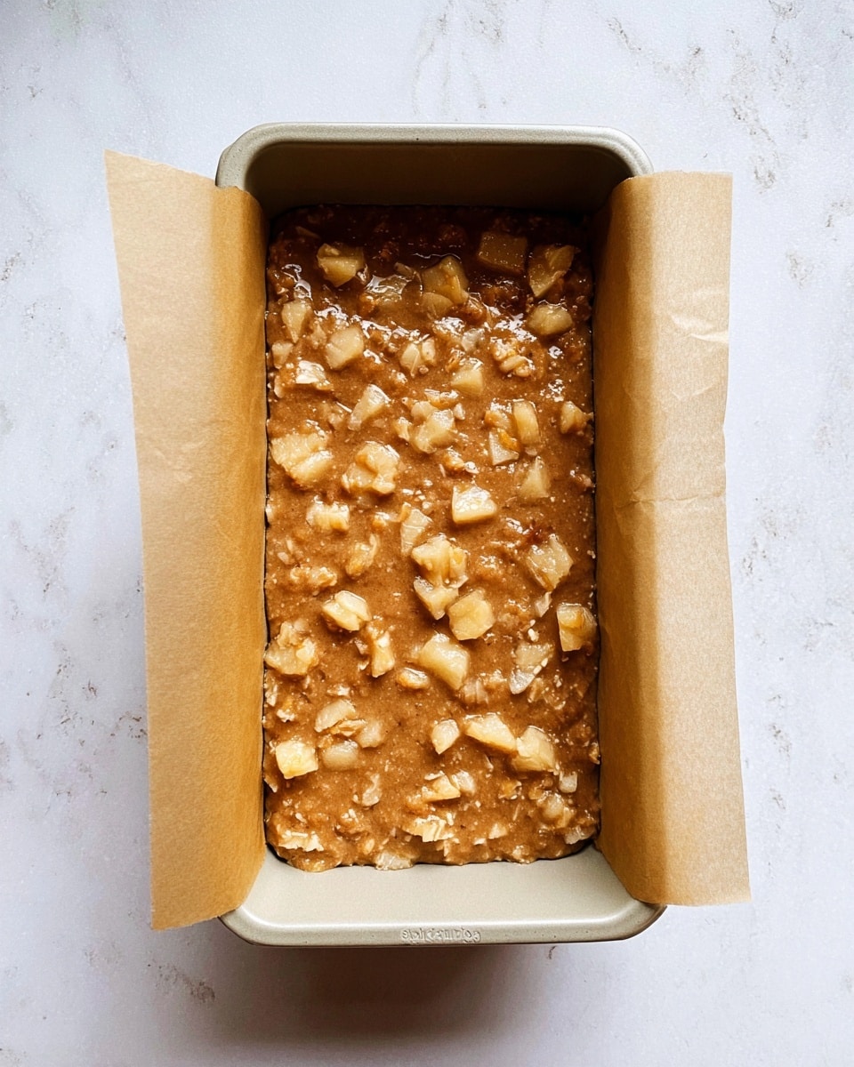A beige rectangular baking pan lined with brown parchment paper filled with a light brown batter mixed with small, uneven chunks of fruit spread evenly across the surface, sitting on a white marbled texture surface photo taken with an iphone --ar 4:5 --v 7
