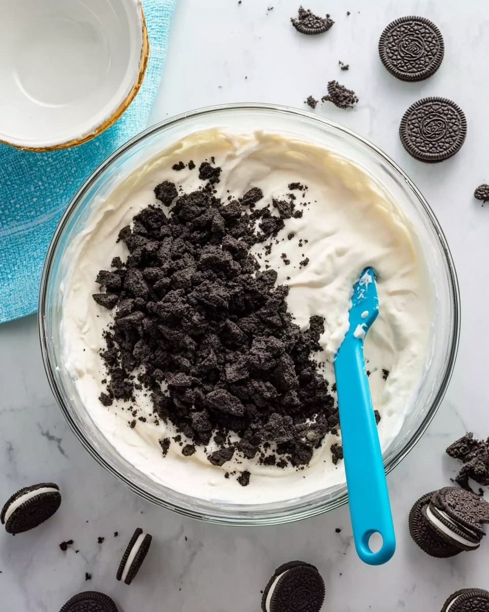 A large clear glass bowl sits on a white marbled surface, filled halfway with smooth white cream. One side of the cream is topped with a thick layer of crushed black and dark brown cookie pieces, which are rough in texture and unevenly spread. A bright blue silicone spatula is placed inside the bowl, resting on the edge where cream and cookies meet. Scattered around the bowl on the white marbled surface are whole and broken sandwich cookies with black outer layers and white filling clearly visible. A small empty white bowl with a gold rim is in the top left corner, and a blue cloth napkin partially peeks in from the left side. Photo taken with an iphone --ar 4:5 --v 7