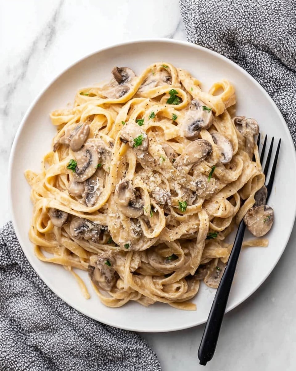 A white plate holds a serving of creamy fettuccine pasta mixed with sliced mushrooms evenly spread throughout. The fettuccine strands are thick and coated with a smooth, light beige sauce. Small green herb pieces are sprinkled on top along with a light dusting of grated cheese or seasoning. A black fork rests on the right side of the plate. The plate sits on a white marbled surface with a grey and white textured cloth nearby. Photo taken with an iphone --ar 4:5 --v 7