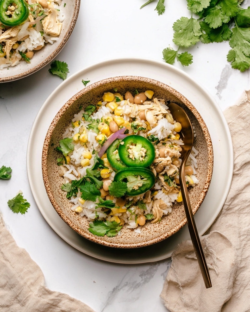 A textured bowl with speckled brown edges sits on a white plate, filled with a mix of white rice, light yellow corn, beige beans, and small diced pale chicken pieces. On top, there are three bright green jalapeno slices, fresh green cilantro leaves, and thin purple onion slices scattered. A bronze spoon rests inside the bowl. The background is a white marbled surface with a beige cloth and scattered cilantro leaves around. Photo taken with an iphone --ar 4:5 --v 7