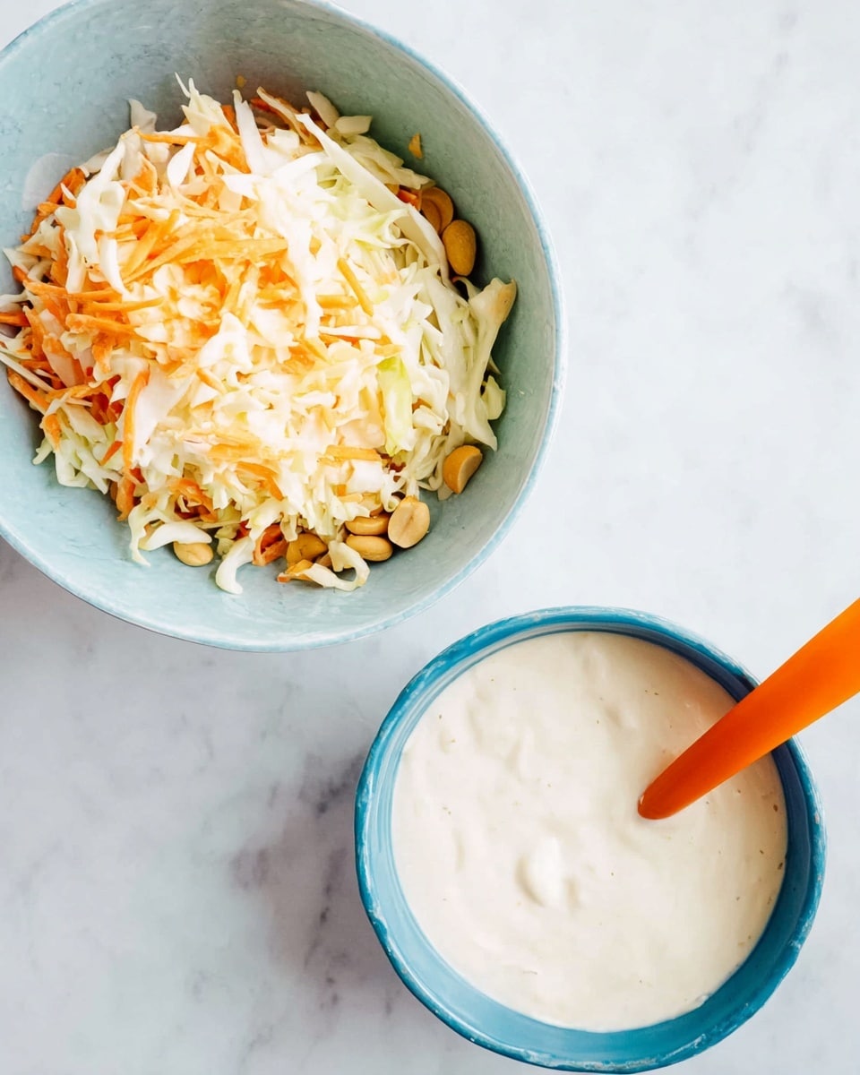 Two light blue bowls are placed on a white marbled surface, one at the top right and one below it. The top bowl is filled with shredded cabbage and carrot, showing mostly white and orange colors mixed together, along with some small peanuts on the right side. The bottom bowl contains a creamy white dressing, smooth and thick, with an orange spoon resting inside, partly covered by the dressing. The scene is bright and clean looking. photo taken with an iphone --ar 4:5 --v 7