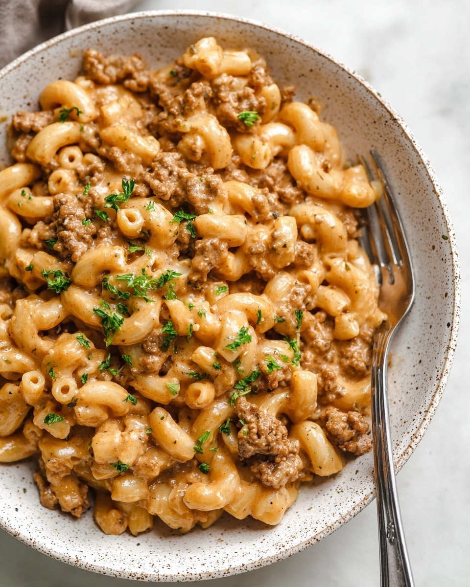 The dish is a close-up of creamy macaroni pasta mixed with ground meat in a white speckled bowl placed on a white marbled surface. The macaroni is covered in a light brown, thick sauce with small pieces of browned ground meat mixed evenly throughout. Fresh green parsley is sprinkled on top, adding a contrast of color. A silver fork rests inside the bowl on the right side. The overall look is hearty and comforting with a smooth texture from the sauce coating the pasta. Photo taken with an iphone --ar 4:5 --v 7
