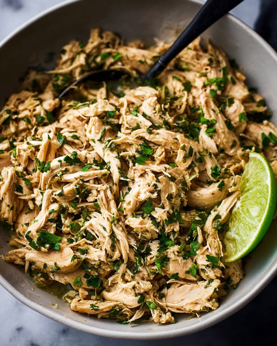 A close-up view of a bowl filled with shredded chicken mixed with chopped green herbs scattered evenly throughout. The chicken pieces are light brown with a moist texture, piled high inside a white bowl. A single lime wedge with bright green skin rests on the edge of the bowl. A dark spoon is partially submerged in the chicken, angled toward the top left of the frame. The background shows a white marbled surface with soft lighting highlighting the textures of the chicken and herbs. Photo taken with an iphone --ar 4:5 --v 7
