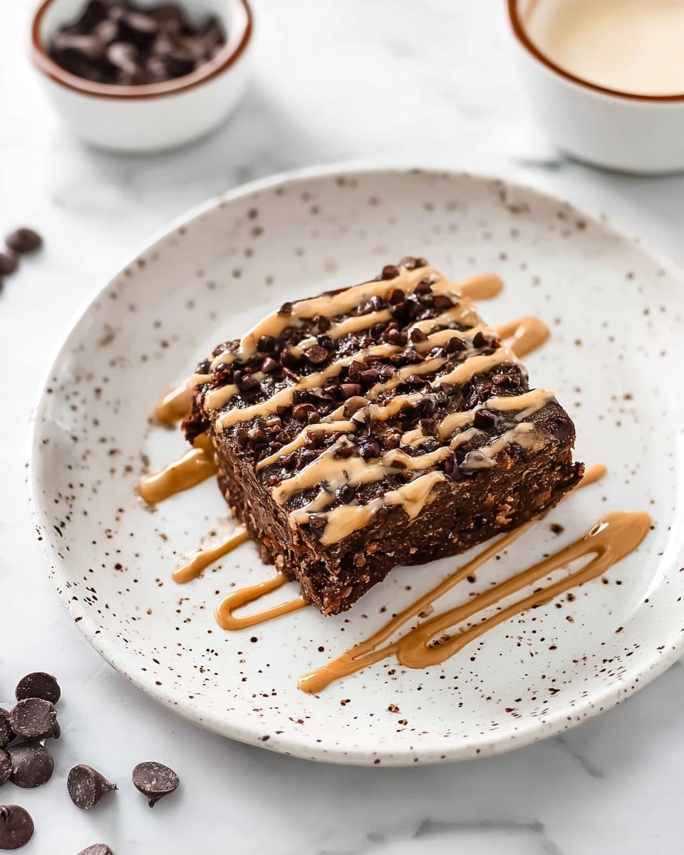 A single dark brown square dessert sits in the middle of a white plate with brown speckles; the dessert looks dense and textured with visible chocolate chips and small bits mixed throughout. The top layer is drizzled with light brown creamy sauce in uneven lines. Around the plate, a few chocolate chips are scattered, and in the background, a small bowl with a white creamy substance and a brown rim is partially visible. The scene is set on a white marbled surface. photo taken with an iphone --ar 4:5 --v 7
