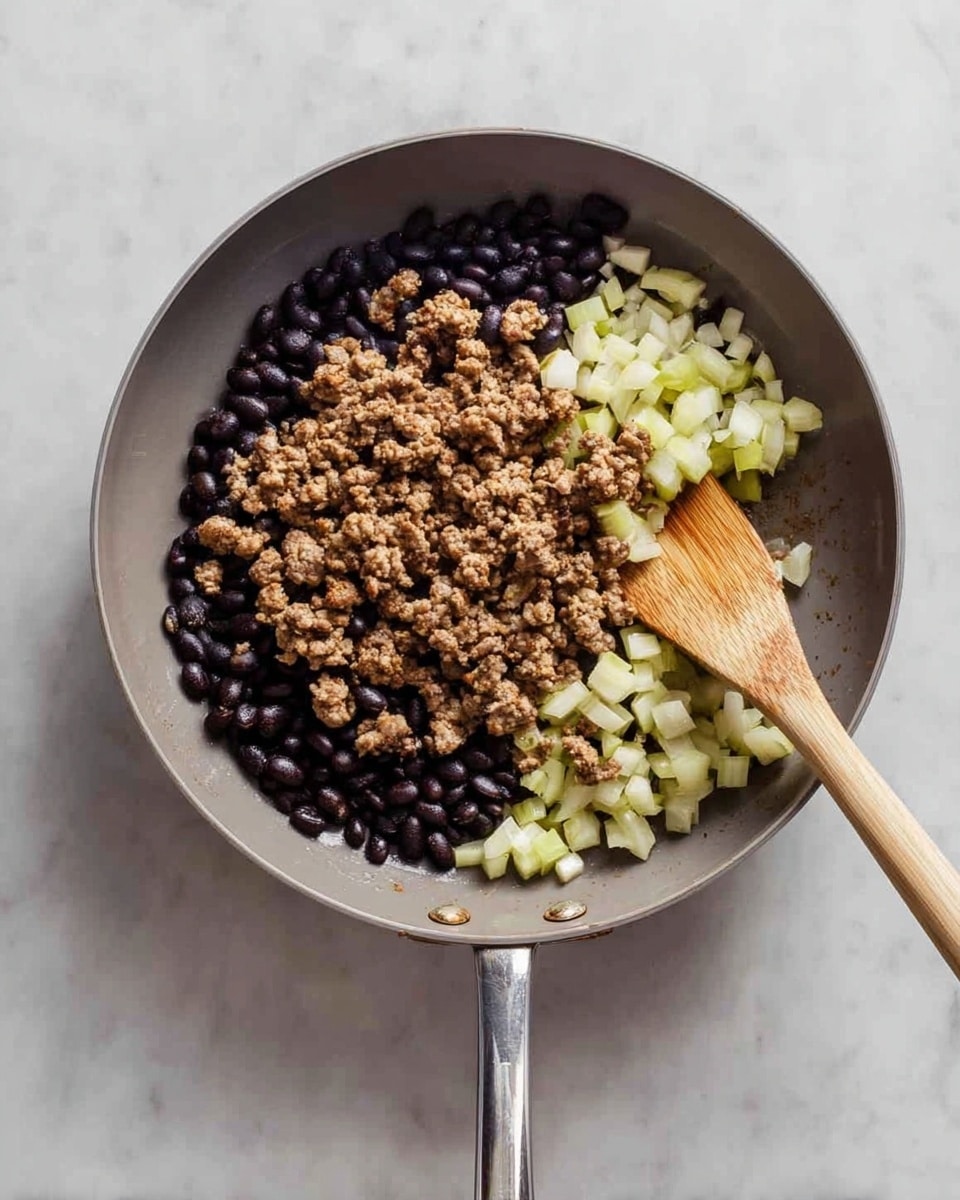 A gray frying pan filled with three main layers of food: a bottom layer of black beans, a middle layer of small pale green and white diced cubes, and a top layer of browned ground meat mixed evenly over the other ingredients. A wooden spoon with a light wooden handle rests inside the pan, partially covered by the food. The pan is placed on a white marbled surface, and the photo is taken from above, showing the entire pan and its long shiny metal handle extending to the right side photo taken with an iphone --ar 4:5 --v 7