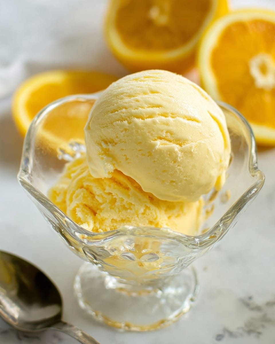 The image shows one round scoop of pale yellow ice cream with a smooth, creamy texture, sitting in a clear, small glass bowl with a decorative, wavy edge. The bowl sits on a white marbled surface. In the blurred background, there are two juicy yellow lemon halves and a silver spoon is partially visible next to the bowl. The light in the photo highlights the cool and soft texture of the ice cream, making it look fresh and inviting. photo taken with an iphone --ar 4:5 --v 7