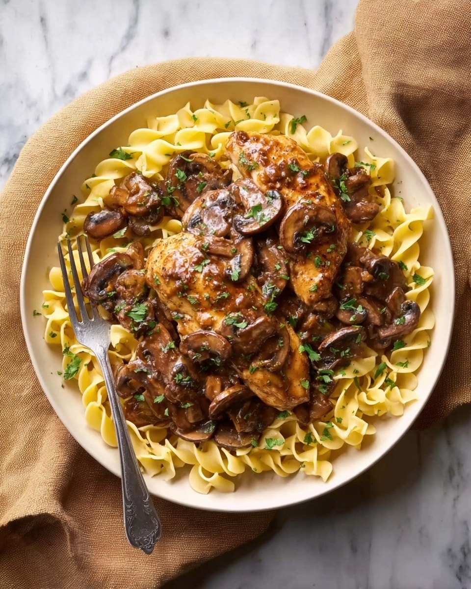 A white plate filled with a bed of yellow egg noodles spread evenly in a circular layer, topped with two pieces of golden-brown cooked chicken covered in a thick, dark brown mushroom sauce with visible sliced mushrooms scattered on and around the chicken. Green chopped herbs are sprinkled on top, adding a touch of color. A silver fork rests on the left edge of the plate, which is placed on a beige cloth on a white marbled surface. photo taken with an iphone --ar 4:5 --v 7