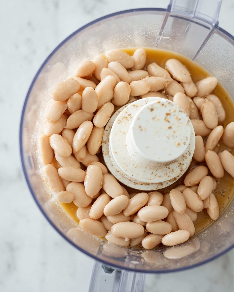 The image shows a close-up of a food processor bowl filled with light beige beans soaking in a light brown liquid. The beans have a smooth texture and are piled around a white central mixing blade with small specks of seasoning visible on it. The food processor bowl is clear plastic, and the background surface is a white marbled texture. photo taken with an iphone --ar 4:5 --v 7