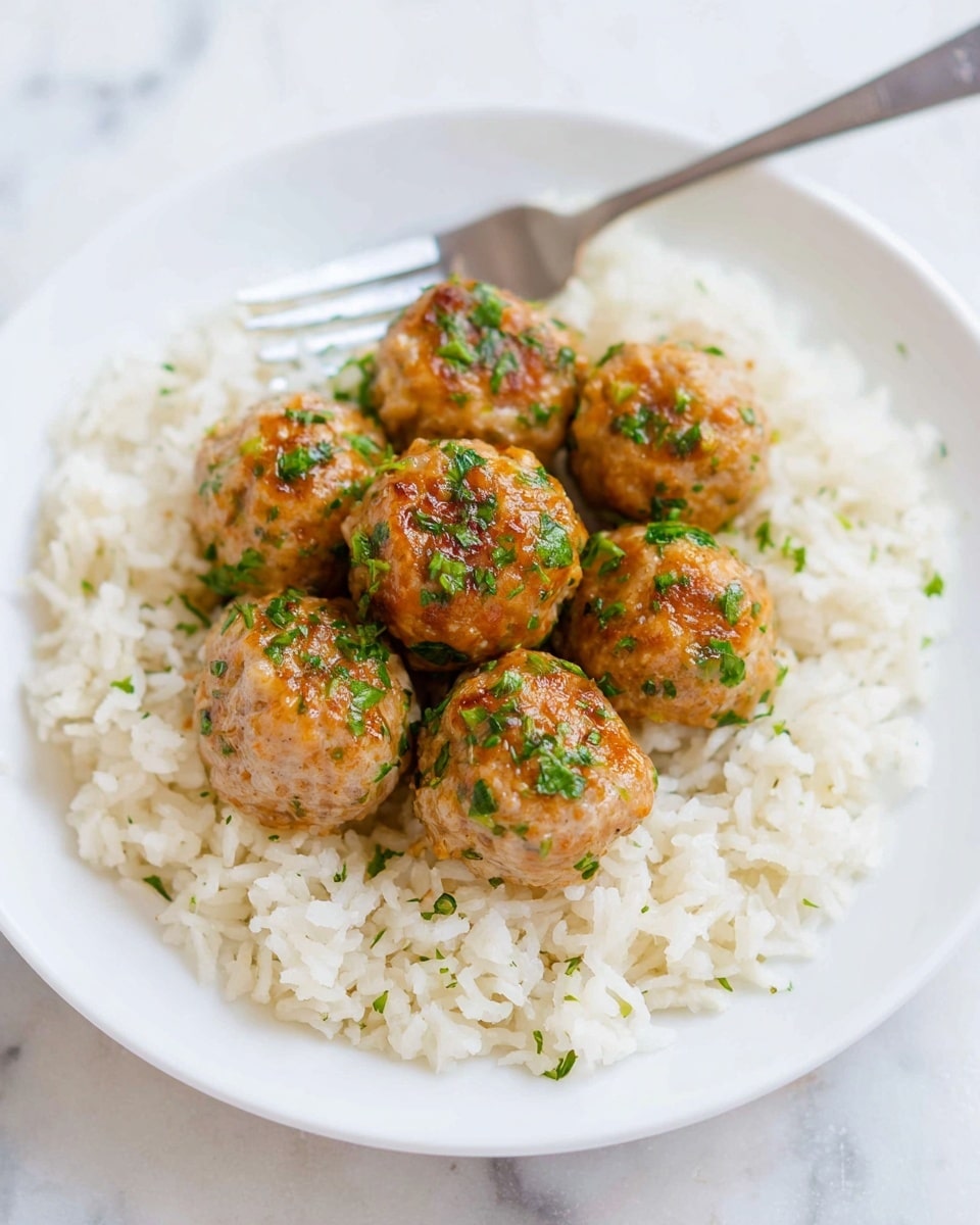 The image shows a white plate with a base layer of fluffy white rice spread evenly in a circular shape. On top of the rice, there are six golden-brown meatballs arranged closely together in a small pile, each meatball speckled with finely chopped green herbs. A silver fork is placed near the top edge of the plate, slightly out of focus. The plate rests on a white marbled surface, adding a clean, simple background that highlights the dish's warm colors photo taken with an iphone --ar 4:5 --v 7