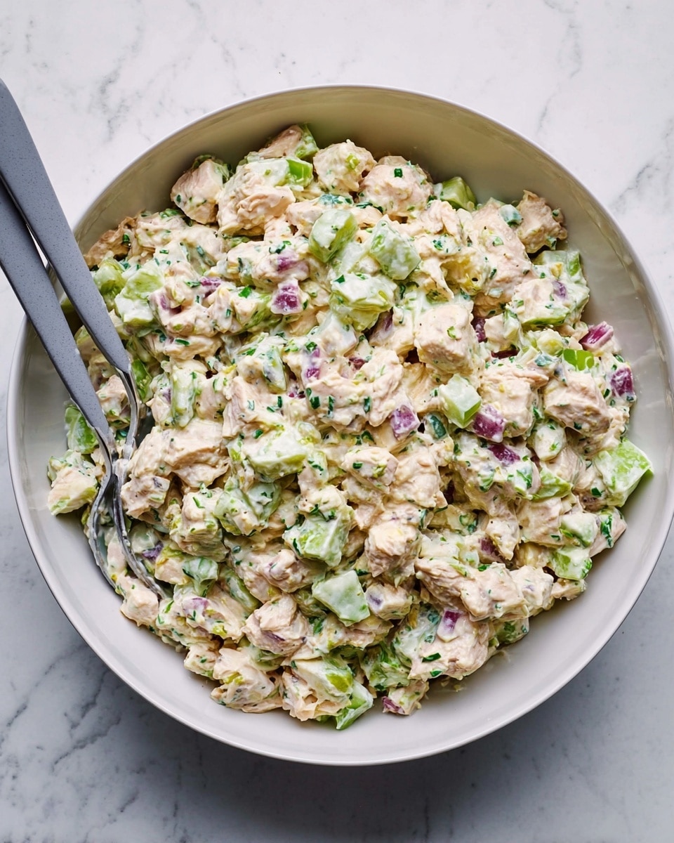 A close-up view of a white bowl filled with a creamy chicken salad mix, showing about three layers of ingredients blended together: chunks of pale pink chicken, finely chopped bright green celery, and small pieces of purple-red onion, all mixed with a white dressing speckled with green herbs. The texture looks chopped and creamy, with visible bits of each ingredient throughout. Two gray-handled spoons rest inside the bowl, one on the left and one on the right. The bowl is placed on a white marbled surface. photo taken with an iphone --ar 4:5 --v 7