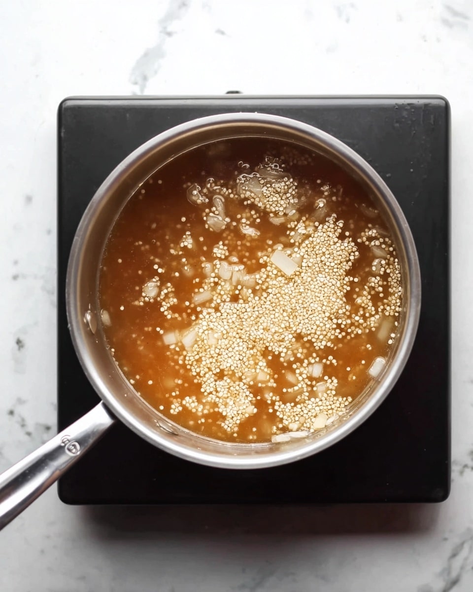 A metal pot sits on a stovetop with light brown liquid inside. The liquid has small white quinoa grains floating on top and some small diced white onion pieces mixed in. The pot is round and shiny with a long handle extending to the left. The stovetop surrounds the pot with a dark edge, and the background surface is a white marbled texture. The image looks simple and clean, showing a cooking process. Photo taken with an iphone --ar 4:5 --v 7