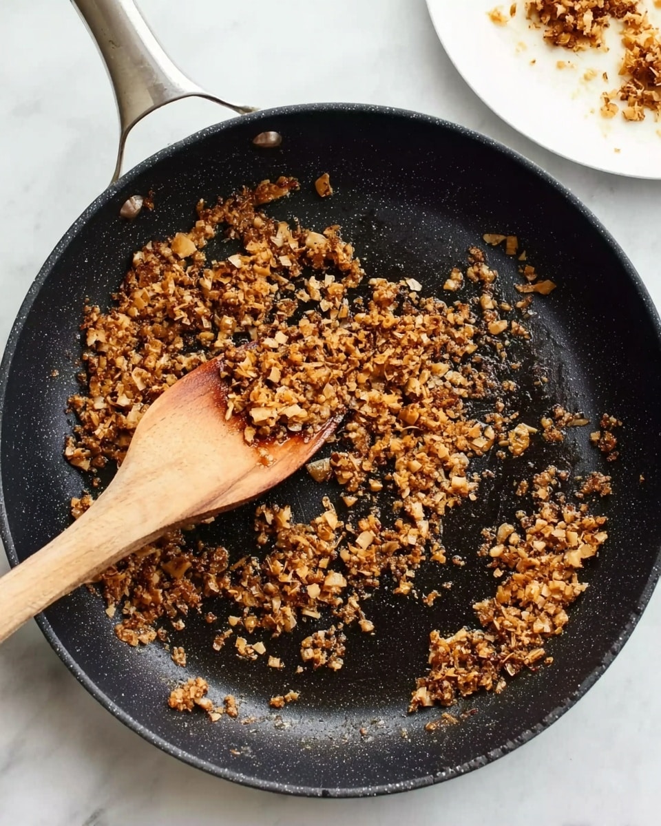 A close-up of a black frying pan on a white marbled surface, filled with small, finely chopped browned pieces of food scattered across the pan's surface. A wooden spoon lies in the pan, partially covered by the browned pieces, its handle extending toward the edge of the pan. The pan handle is visible at the top left corner, and there is a partial view of a white plate with food on the top right side of the image. photo taken with an iphone --ar 4:5 --v 7