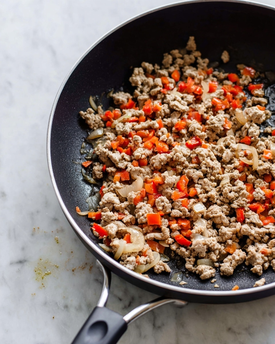 A close-up view of a black cooking pan with a small amount of cooked ground meat mixed with small diced red bell peppers and translucent cooked onions spread loosely inside the pan, showing a mix of light brown, orange-red, and pale yellow colors. The pan has black handles and sits on a white marbled surface, with small bits of food and splashes visible on the inner side of the pan. The lighting is soft, highlighting the texture of the cooked ingredients and the smooth surface of the pan. photo taken with an iphone --ar 4:5 --v 7