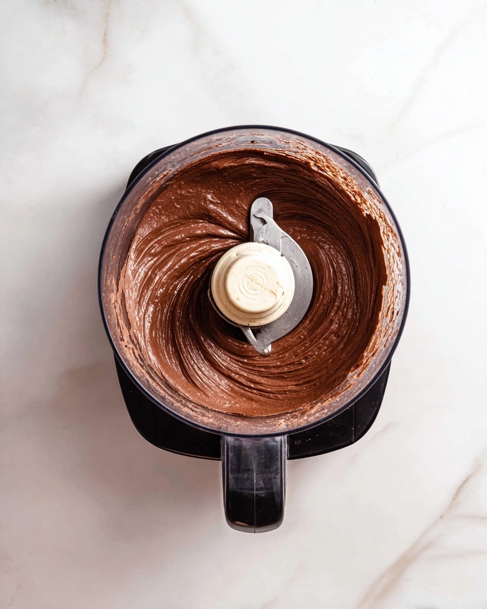 A top view of a black food processor with a clear mixing bowl on a white marbled surface, filled with smooth, creamy, dark brown chocolate mixture. The texture is thick and well blended, clinging evenly to the sides of the bowl and swirling around the silver blade in the center. Photo taken with an iphone --ar 4:5 --v 7