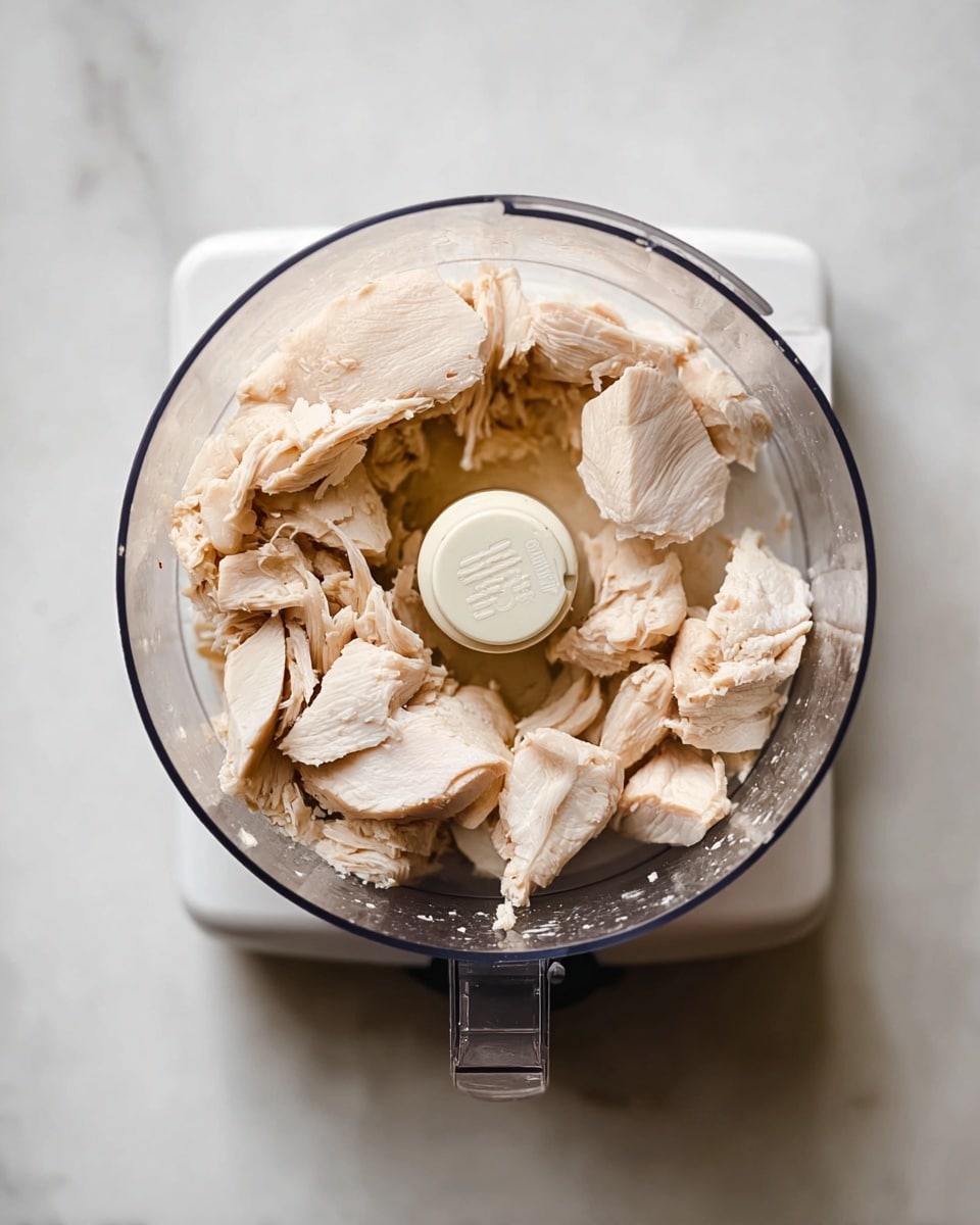 The image shows a top view of a food processor bowl filled with several pieces of light beige cooked chicken placed irregularly around the center blade. The inside of the bowl is clear, showing the smooth texture of the chicken, with some pieces torn and soft-looking. The food processor base is white and sits on a white marbled surface. The overall look is clean and simple, with natural light highlighting the soft colors of the chicken and plastic bowl. Photo taken with an iphone --ar 4:5 --v 7