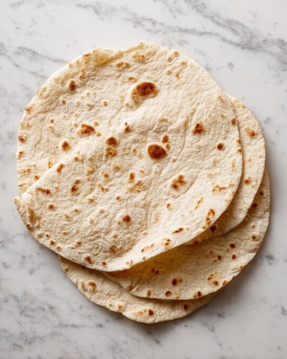 The image shows three round flatbreads stacked slightly overlapping on a white marbled surface. Each flatbread is light tan with small, scattered brown spots and slight puffed areas, giving a soft and airy texture. The edges are slightly uneven, showing a handmade look. The flatbreads are thin and quite large, and their simple color and texture stand out against the white background. photo taken with an iphone --ar 4:5 --v 7