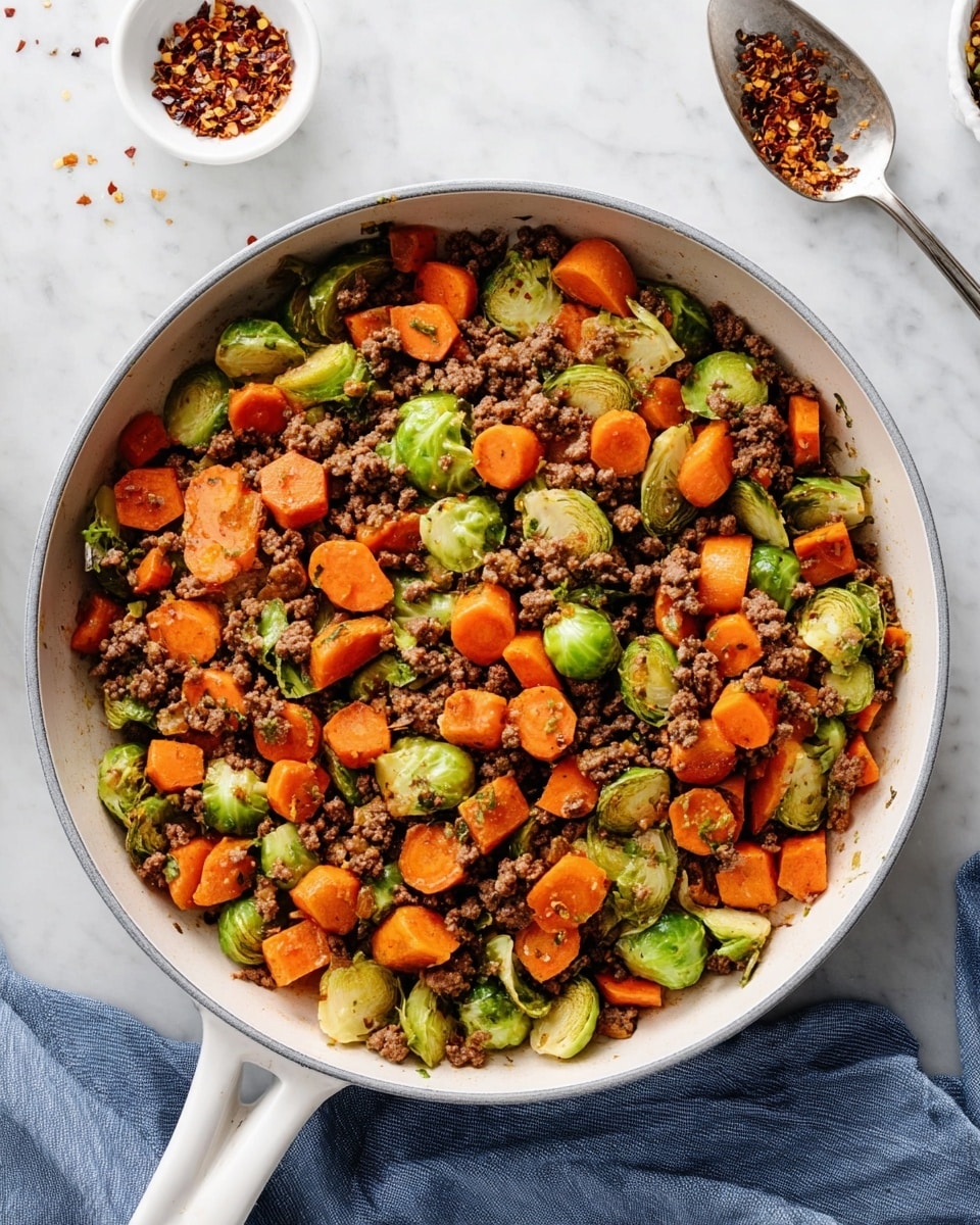 A white pan filled with three main layers mixed together: small, round orange carrot slices; small green Brussels sprout halves with a slightly glossy look; and browned ground beef bits with a crumbly texture. The vegetables and meat are evenly spread and mixed, showing a balance of bright green, vibrant orange, and rich brown colors with black pepper specks on top. The pan sits on a white marbled surface with a steel spoon containing red pepper flakes above it, a small white bowl with cracked chili flakes to the left, and a blue cloth napkin to the right. Photo taken with an iphone --ar 4:5 --v 7