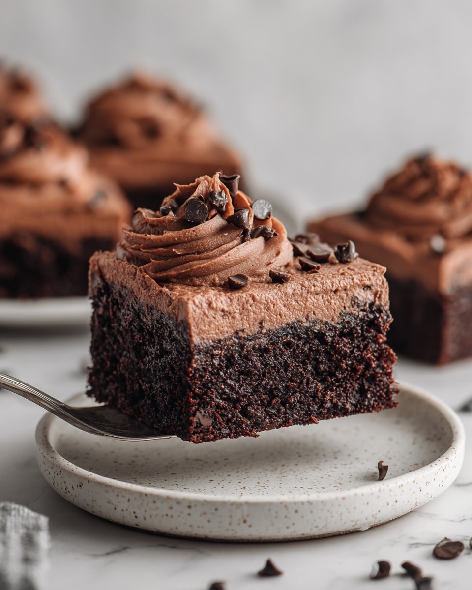 The image shows a close-up of a rich chocolate brownie square with two main layers: a dense dark brown base with a slightly crumbly texture and a thick, creamy chocolate frosting layer on top. The frosting is swirled and decorated with small, shiny chocolate chips scattered across the surface. This brownie square sits on a white speckled plate, and in the background, there are more cut brownie pieces. A silver spatula is lifting the brownie, and the setting includes a white marbled surface. photo taken with an iphone --ar 4:5 --v 7