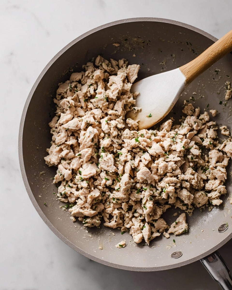 A close-up view of a gray non-stick frying pan filled with cooked ground chicken mixed with small bits of green herbs. The cooked chicken is light beige with some parts slightly darker, showing a soft texture. In the center, a white spoon with a wooden handle scoops up a portion of the chicken, mixing it gently in the pan. The pan rests on a surface with white marbled texture, and no other objects are visible in the frame. photo taken with an iphone --ar 4:5 --v 7