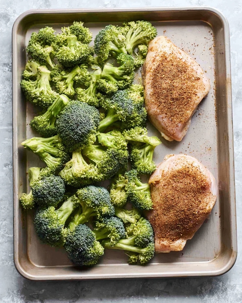 A light gray baking sheet holds two large raw chicken pieces at the top right corner, each coated with a brown spice mix, creating a textured layer on the pale chicken skin. Below the chicken, on the left side of the sheet, a generous pile of fresh broccoli florets spreads out, their vibrant green color contrasting with the smooth gray surface. The broccoli pieces vary in size, showing detailed bumpy textures on the crown and crisp smooth stems. The overall look is clean and fresh on a white marbled textured surface. Photo taken with an iphone --ar 4:5 --v 7