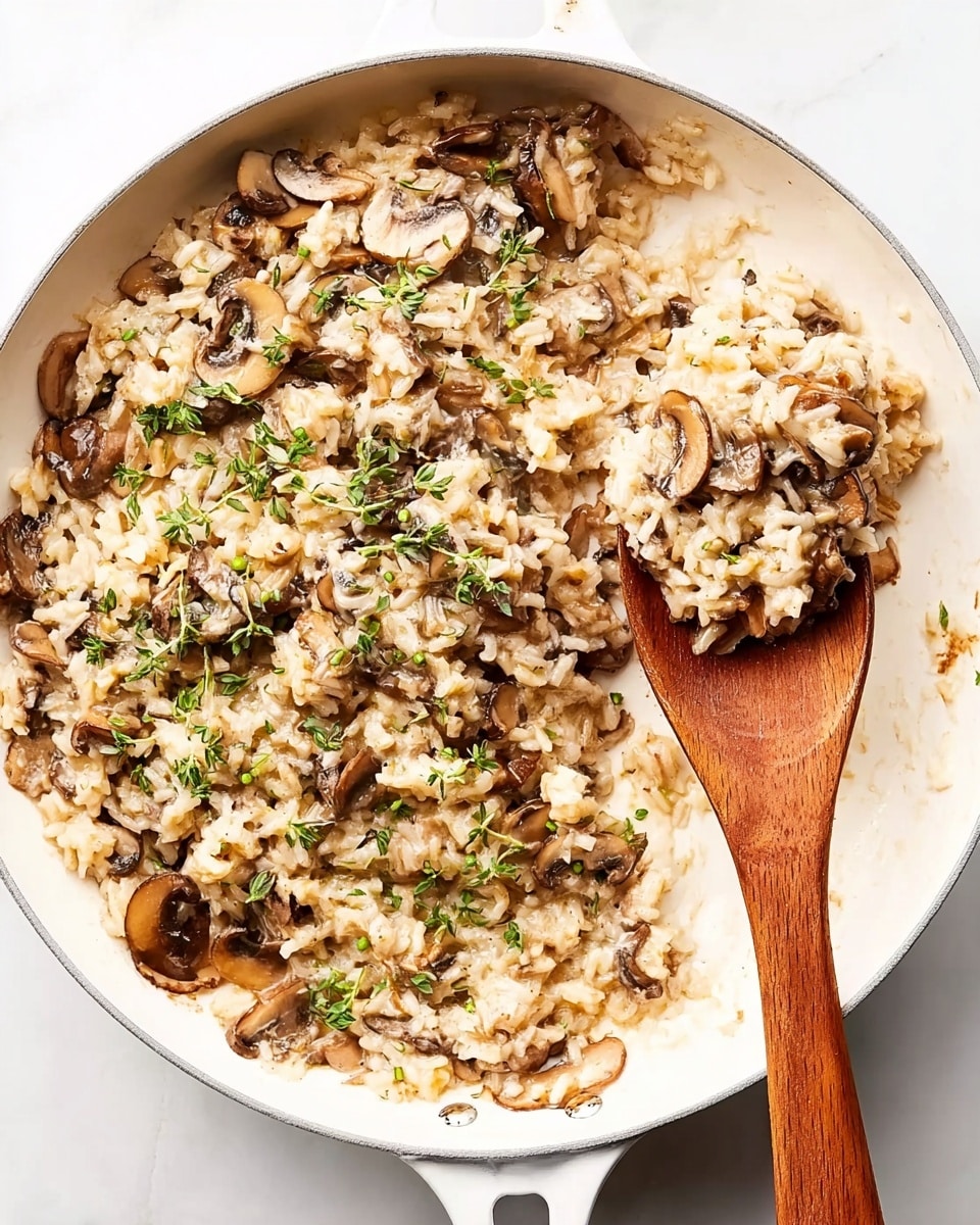 A white frying pan filled with a creamy mushroom dish that has one visible layer, showing small pieces of soft rice mixed with thinly sliced brown mushrooms and green herb sprigs sprinkled on top. A wooden spoon inside the pan scoops a portion, lifting mushrooms and rice, revealing a light tan and brown mix with textures from the soft rice and tender mushrooms. The pan rests on a white marbled surface. photo taken with an iphone --ar 4:5 --v 7