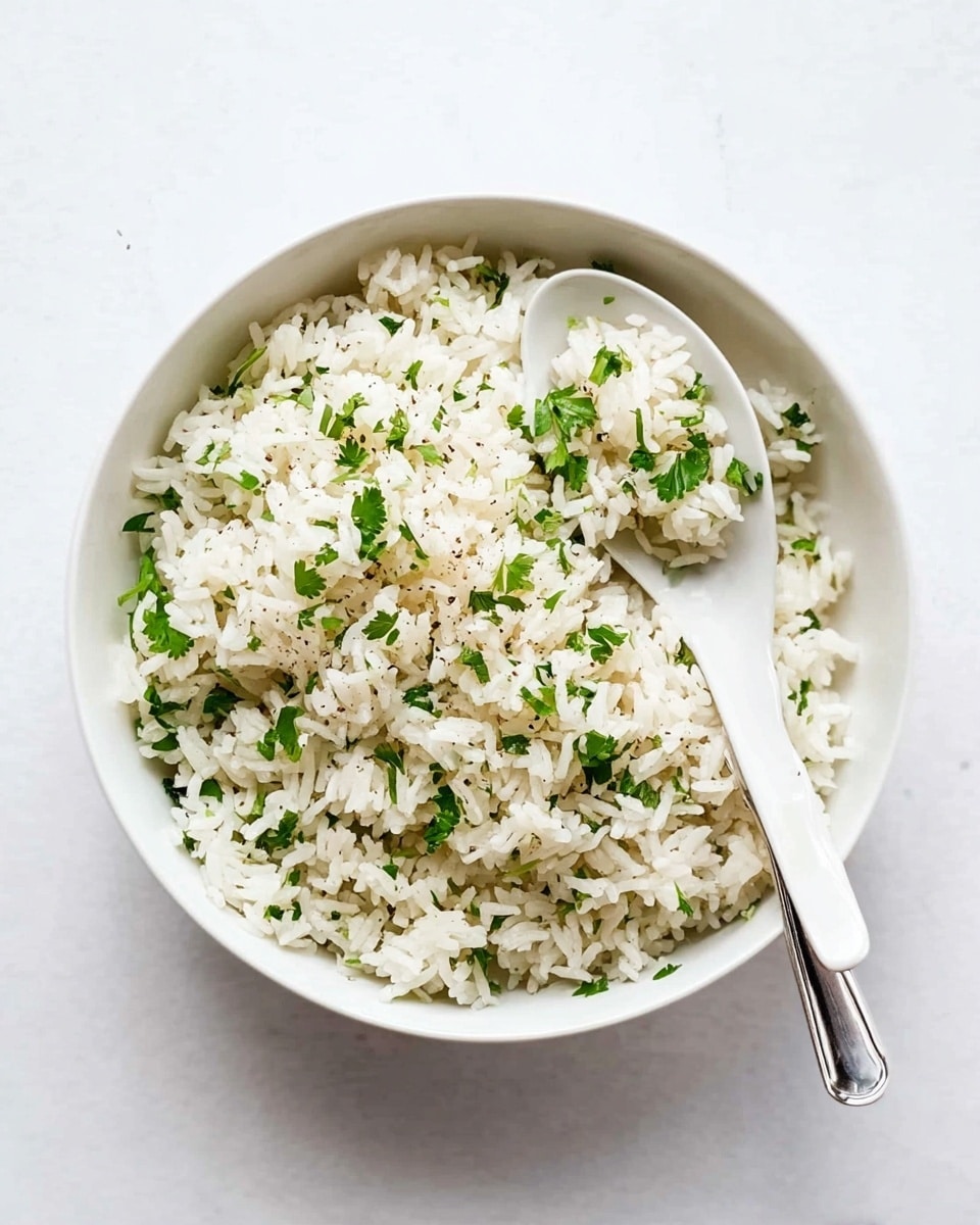 A white bowl filled with cooked white rice mixed evenly with small green parsley leaves, giving a fresh look. The rice grains look soft and fluffy with some black pepper specks sprinkled throughout. Inside the bowl, a white ceramic spoon rests on the rice with a small scoop of the rice and parsley mix on it. The bowl sits on a white marbled surface, and the overall colors are mostly white with vibrant green from the parsley, creating a simple and clean presentation. photo taken with an iphone --ar 4:5 --v 7