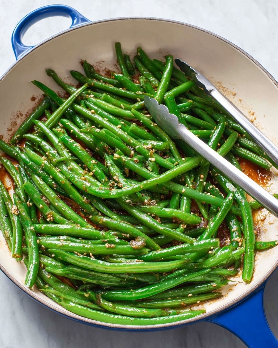 The image shows a white pan with a blue handle filled with cooked green beans. The beans are bright green and shiny, spread evenly across the pan, with some light browning on a few beans. There are small bits of garlic and sesame seeds scattered over the beans, adding texture and detail. A pair of metal tongs rests on the right side of the pan. The pan sits on a white marbled surface. photo taken with an iphone --ar 4:5 --v 7