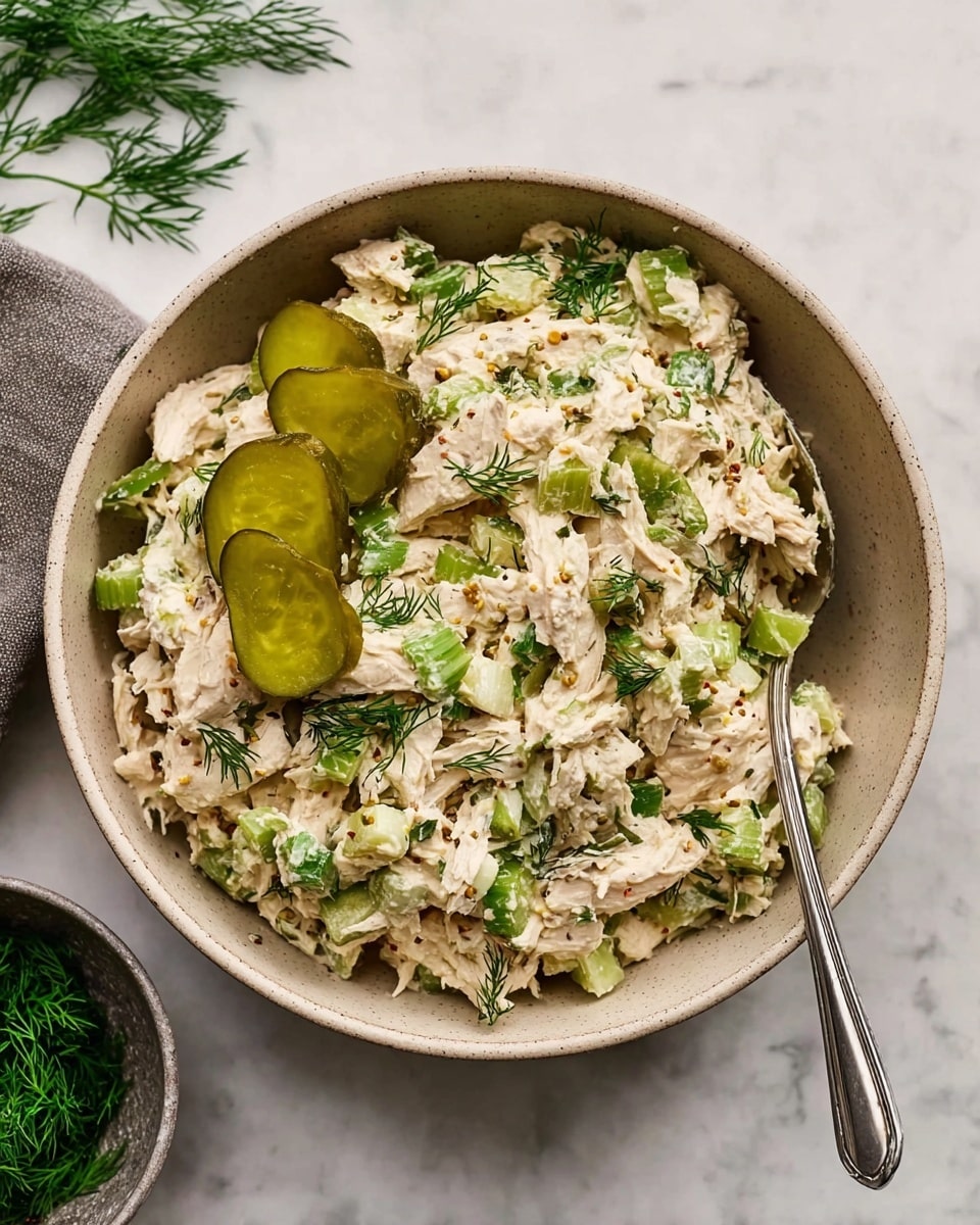A beige bowl filled with creamy chicken salad made of shredded white chicken mixed with chopped green celery and fresh green dill, all coated in a light mayo-like dressing with visible mustard seeds. On top are two slices of green pickle and more dill sprinkled over the whole dish, giving a fresh touch. A silver spoon rests inside the bowl on a white marbled surface with a few dill sprigs nearby and a small bowl of fresh dill in the corner. photo taken with an iphone --ar 4:5 --v 7