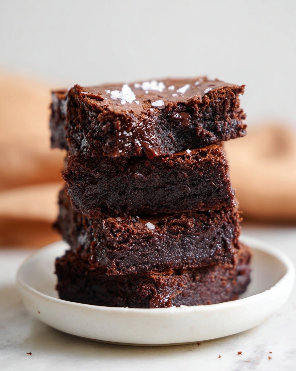 A stack of four brownies sits on a white plate on a white marbled surface. Each brownie is thick and dark brown with a rich, crumbly texture. The top brownie is slightly broken at the corner, showing a moist inside. Small flakes of white salt are scattered on the brownies. The background is softly blurred with warm tones, focusing all attention on the brownies. photo taken with an iphone --ar 4:5 --v 7
