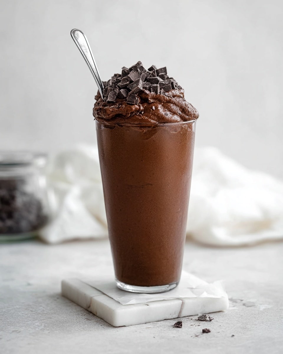 A tall clear glass filled with a smooth dark chocolate pudding layer at the bottom, topped with a thick, rough textured dark chocolate whipped cream layer topped with chocolate chunks, with a silver spoon inserted in the glass. The glass stands on a small white marbled tile that slightly overlaps a sheet of white paper. The background and surface have a clean white marbled texture. A blurred jar and folded white cloth are faintly visible in the back. Photo taken with an iphone --ar 4:5 --v 7