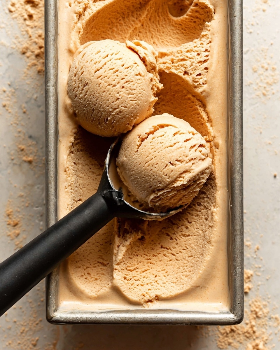 A close-up image shows two large scoops of light brown creamy ice cream with a slightly rough texture resting on top of a full rectangular silver metal container filled with the same ice cream. One scoop is held by a black ice cream scooper that lies diagonally across the container, while the other scoop is just above the surface. The ice cream looks soft and has a swirled, slightly airy surface in the container. The background includes a white marbled texture. Photo taken with an iphone --ar 4:5 --v 7