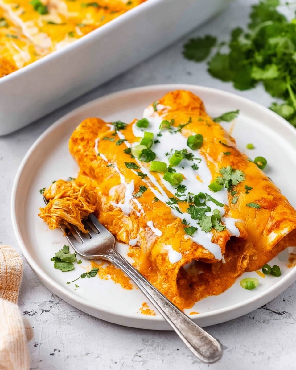 A white round plate holds a single rolled enchilada covered in bright orange sauce with melted cheese on top. The enchilada is garnished with chopped green onions and fresh cilantro leaves scattered over a drizzle of white sauce. A fork with some shredded chicken-like filling is placed on the left side of the enchilada. The plate sits on a white marbled surface with a white baking dish filled with more enchiladas partially seen in the top right corner and a bunch of fresh cilantro at the bottom right. The colors are warm and vibrant, highlighting the creamy, saucy texture of the dish. Photo taken with an iphone --ar 4:5 --v 7