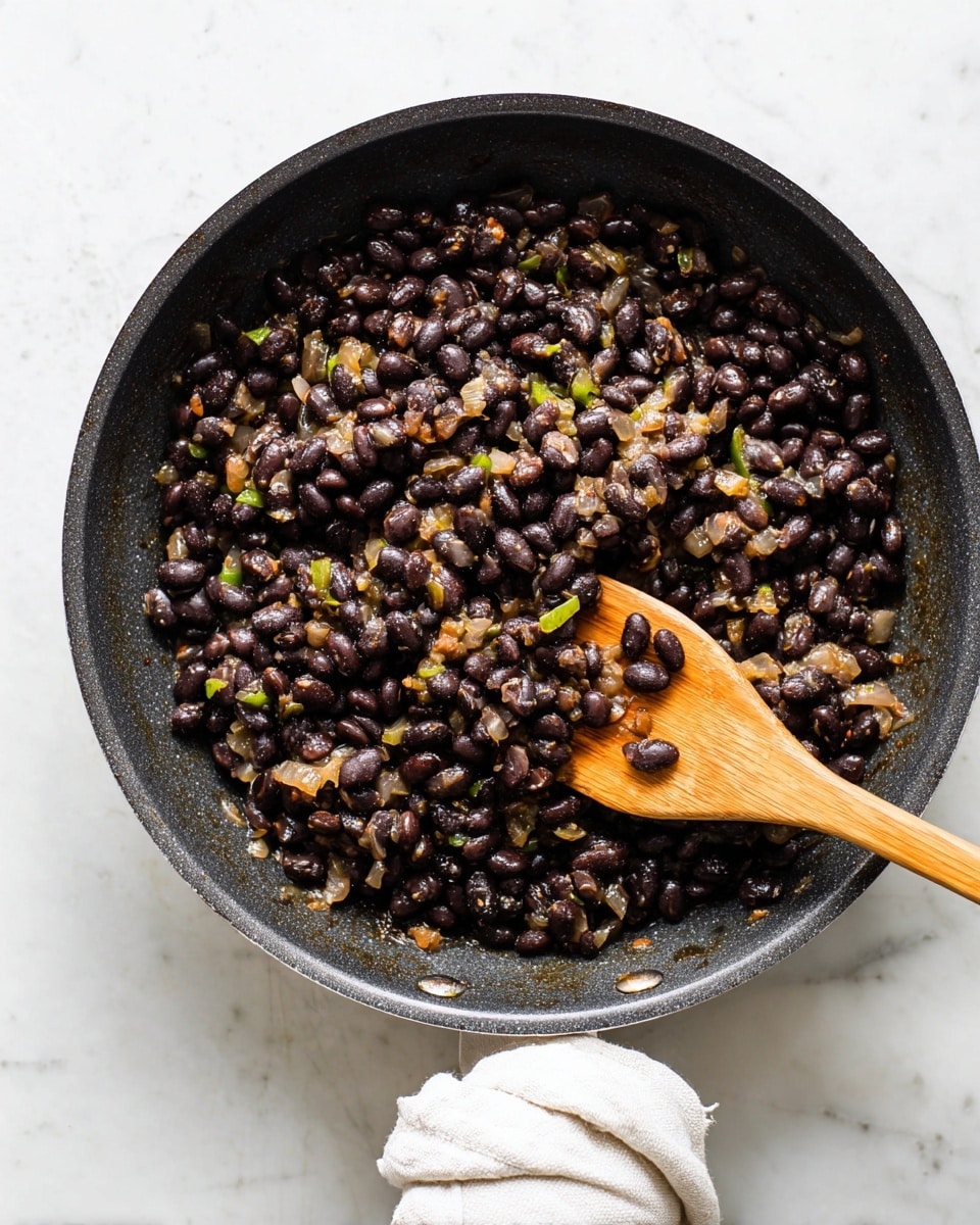 A black pan filled with cooked black beans mixed with small pieces of sautéed onions and green peppers, showing a slightly oily texture that coats the beans and vegetables evenly. A light brown wooden spatula rests inside the pan on the right side, partially covered with some beans. The pan is sitting on a surface with a white marbled texture, and below the pan, a white cloth is visible, wrapped around its handle. photo taken with an iphone --ar 4:5 --v 7