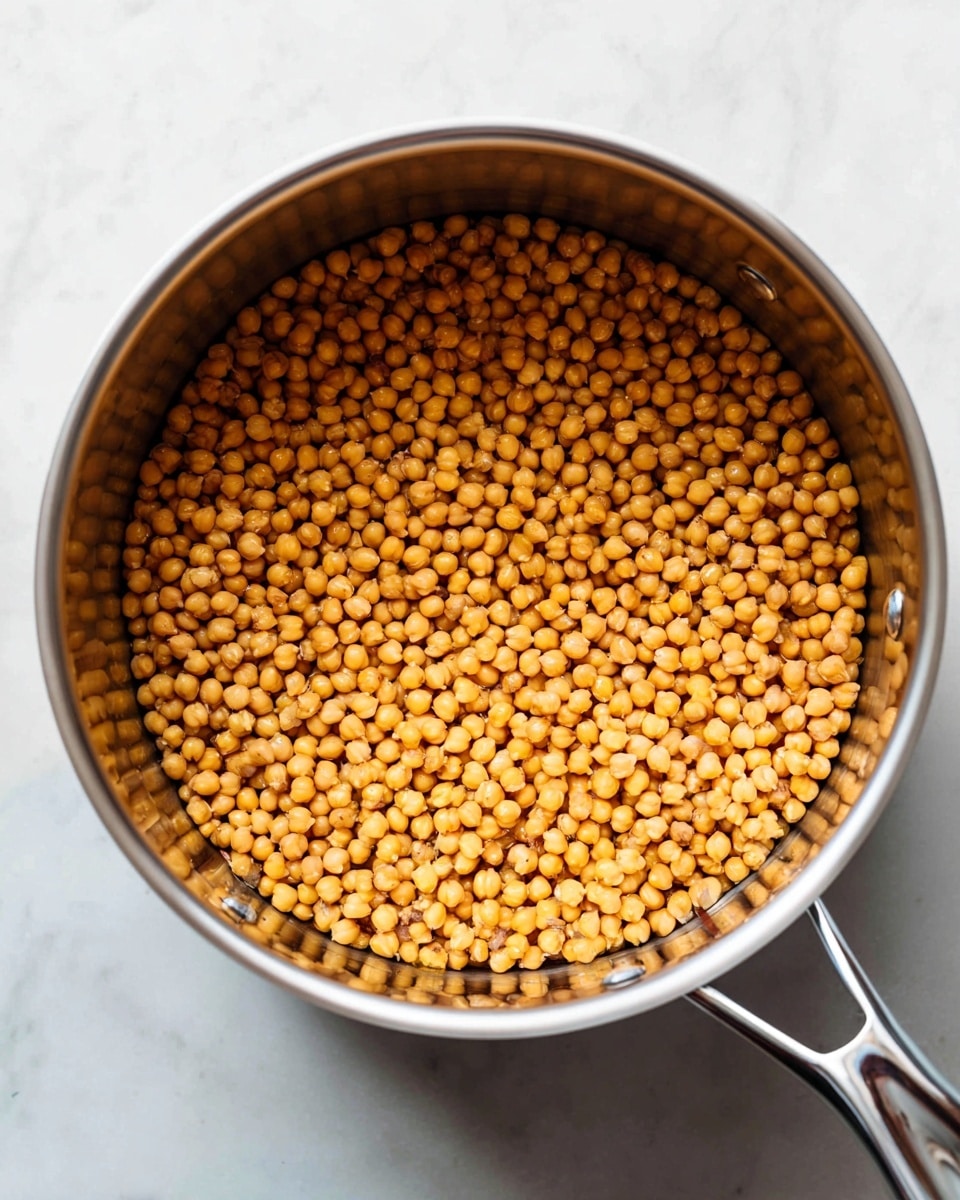 The image shows a close-up top view of a single layer of small, round, golden-brown chickpeas spread evenly inside a large metal pot. The pot has smooth silver walls with two small rivets visible on the side, and a shiny metal handle partially in the frame. The background is a white marbled surface that contrasts with the warm tones of the chickpeas. The light creates a soft shadow inside the pot, highlighting the texture and slight color variations of the chickpeas. Photo taken with an iphone --ar 4:5 --v 7