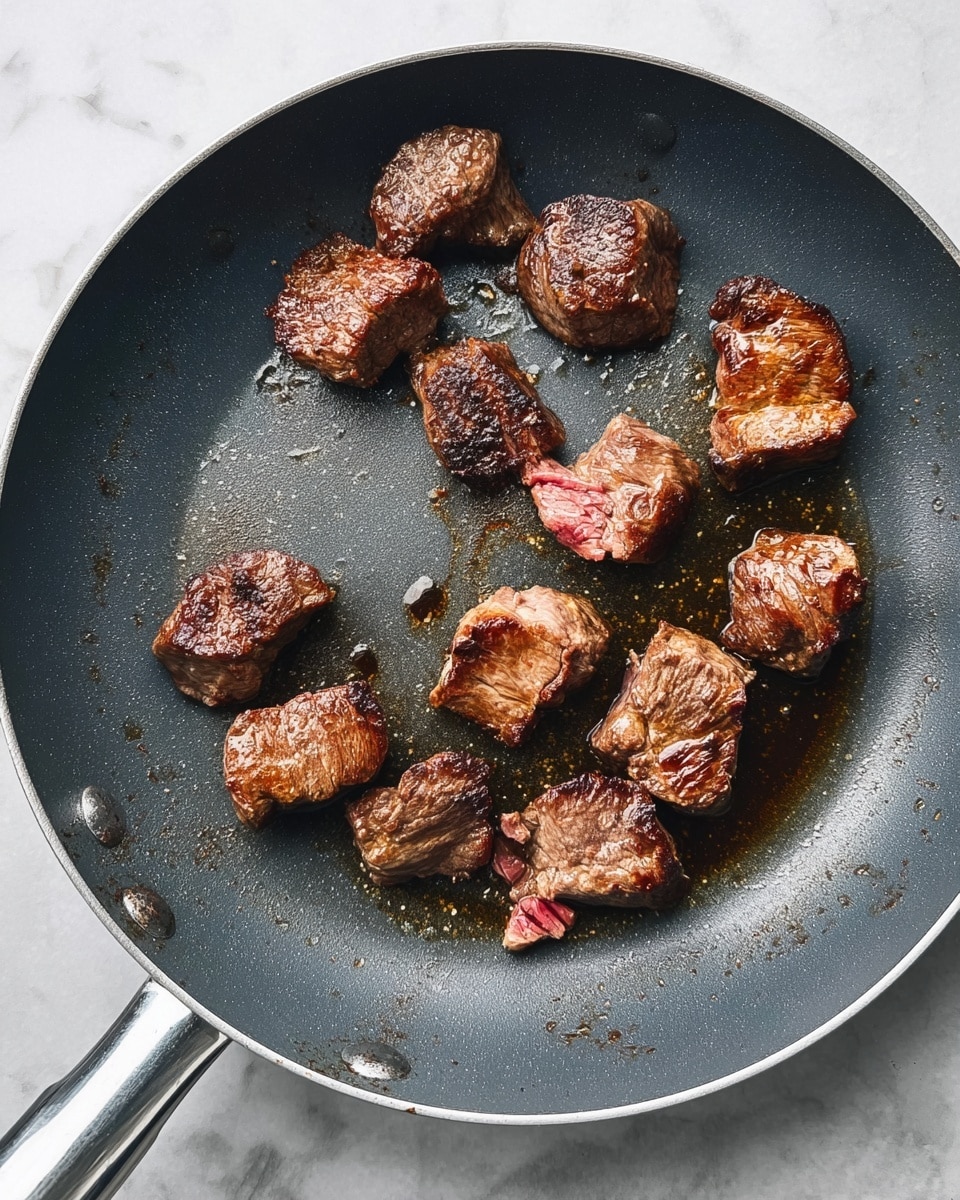 A close-up view of a dark grey frying pan with about eleven small pieces of browned meat scattered around the bottom. Each piece shows a crusty, dark brown outer layer with some parts still showing a reddish raw inside. The meat edges are irregular, and there's a light shine of cooking oil pooling around them on the pan's smooth surface. The pan handle is silver and visible on the left, resting on a white marbled texture background. photo taken with an iphone --ar 4:5 --v 7