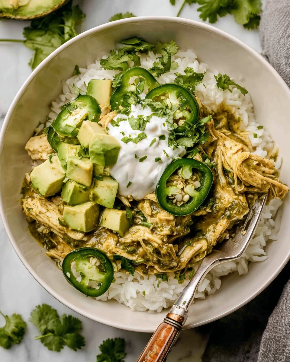 A white bowl on a white marbled surface holds a dish with three main layers: the bottom layer is white rice, fluffy and spread evenly; the middle layer is shredded cooked chicken mixed with a green, creamy sauce containing herbs and spices; the top layer includes fresh green jalapeño slices arranged on the rice edges, chopped avocado cubes, a dollop of white sour cream in the center, and sprigs of bright green cilantro scattered on top and around the bowl. A fork with a wooden handle rests inside the bowl, touching the chicken and rice. Photo taken with an iphone --ar 4:5 --v 7