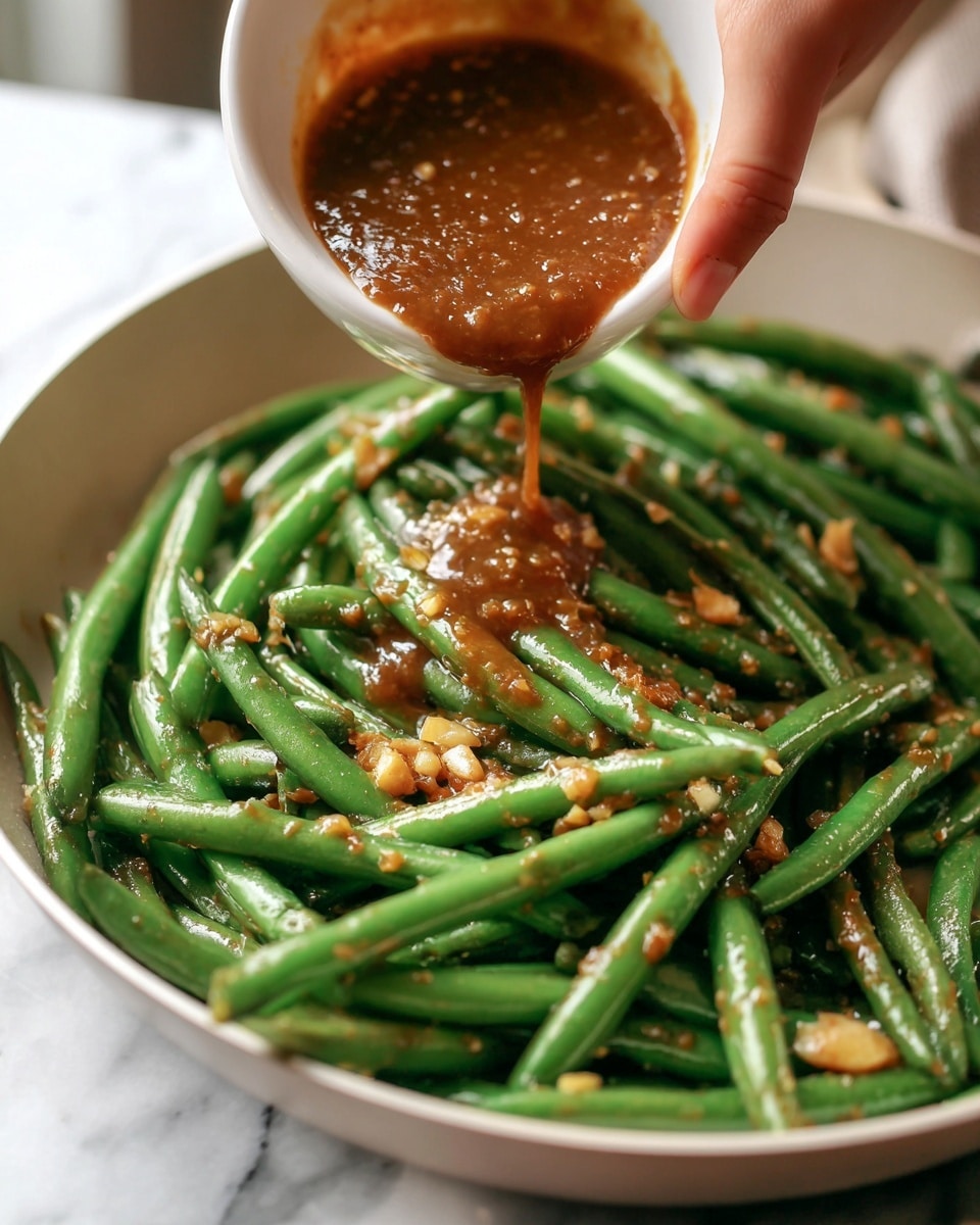 A close-up image shows bright green cooked green beans inside a white pan. A white bowl is held above the pan by a woman's hand, pouring a thick brown sauce onto the green beans. The green beans have a shiny texture and are mixed with small bits of light-colored garlic. The background has a white marbled texture. photo taken with an iphone --ar 4:5 --v 7