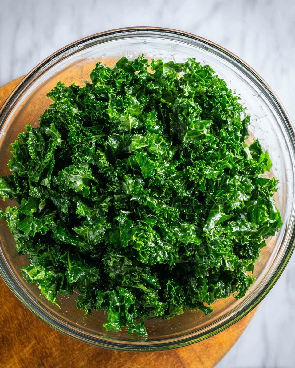 A large clear glass bowl filled with many pieces of chopped kale. The kale is bright green with a shiny, wet look showing the texture of the curly leaves. The bowl sits on a smooth white marbled surface that shines under the light. The kale fills the bowl well, with a mix of small and medium pieces all piled up closely. photo taken with an iphone --ar 4:5 --v 7