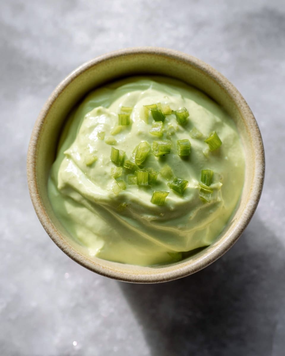 A close-up top view of a small beige ceramic bowl filled with smooth, light green creamy sauce, slightly whipped with soft peaks and gentle swirls. The sauce is garnished with small, bright green diced pieces sprinkled lightly on top. The bowl is placed on a white marbled surface with soft light reflections. Photo taken with an iphone --ar 4:5 --v 7