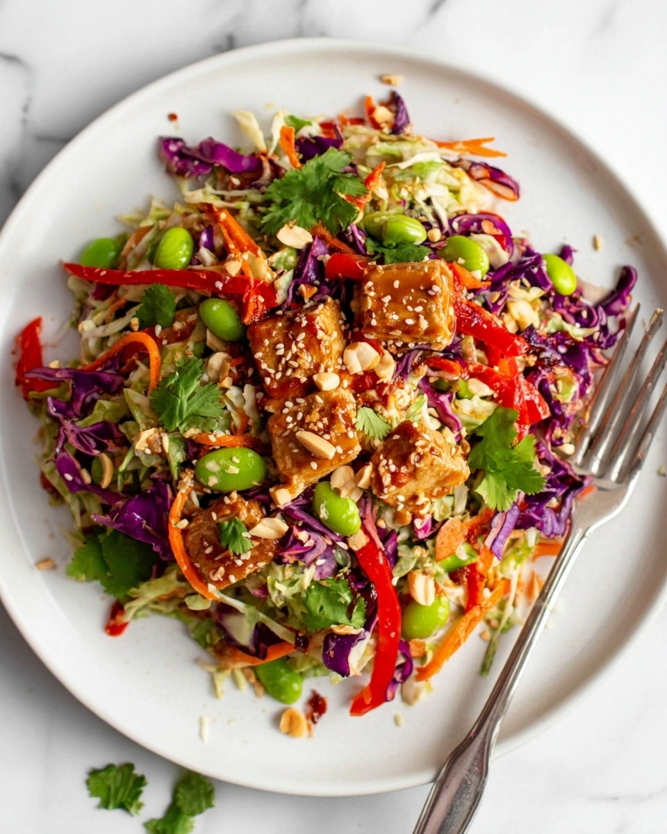 A white plate sits on a white marbled surface, filled with a colorful layered salad. The bottom layer is shredded light green cabbage mixed with shredded purple cabbage, creating a textured and leafy base. Scattered on top are thin strips of bright red bell pepper and small orange carrot pieces, adding vibrant color and crunch. There are green edamame beans spread throughout, along with chunks of brown sauce-coated tofu placed on the salad. The dish is garnished with sesame seeds, chopped peanuts, and fresh green cilantro leaves, giving a nutty and fresh touch. A silver fork rests on the right edge of the plate. Photo taken with an iphone --ar 4:5 --v 7
